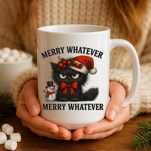 Puede incluir: Taza de cerámica blanca con un gato negro con gorro de Papá Noel, un muñeco de nieve y el texto "MERRY WHATEVER". La taza se sostiene con ambas manos.