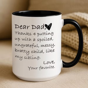 Puede incluir: Taza de cerámica blanca con interior y asa negros. La taza presenta el texto "Dear Dad" seguido de un mensaje humorístico sobre el hermano o hermana de un niño. La taza es un regalo para un padre.