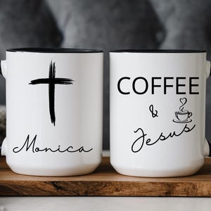 Puede incluir: Dos tazas de cerámica blancas con asas y bordes negros. Una taza presenta una cruz negra y el nombre "Monica". La otra taza tiene las palabras "COFFEE & Jesus" con una ilustración de taza de café.