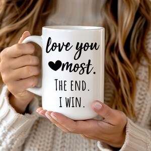 Puede incluir: Taza de cerámica blanca con el texto "love you most. The end. I win." en escritura negra. La taza tiene un asa grande y la sostiene una persona que lleva un suéter color crema.