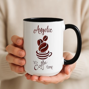 Puede incluir: Taza de café blanca con interior y asa negros. La taza presenta la palabra "Angelic" sobre un diseño de granos de café y una taza. La frase "It's coffee time" está escrita debajo del diseño de la taza.