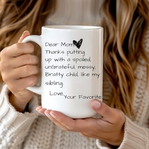 Puede incluir: Taza de cerámica blanca con texto negro que dice: "Querida mamá, Gracias por aguantar a un niño mimado, ingrato, desordenado, malcriado, como mi hermano/a. Con amor, tu favorito/a" y un corazón negro.