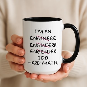 Puede incluir: Taza de cerámica blanca con interior y asa negros. La taza presenta el texto "I'M AN ENGINERE ENGINIERR ENGENIER I DO HARD MATH." La palabra "engineer" está tachada varias veces.