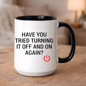 Puede incluir: Taza de cerámica blanca con interior y asa negros. La taza presenta el texto "HAVE YOU TRIED TURNING IT OFF AND ON AGAIN?" en negro, con un icono rojo de botón de encendido. La taza está sobre una superficie de madera.