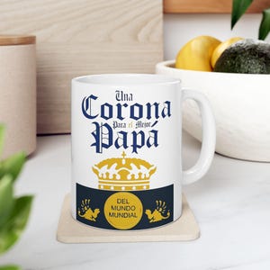 Puede incluir: Taza de cerámica blanca con un diseño azul y dorado. La taza presenta el texto "Una Corona Para el Mejor Papa" y una corona dorada. La parte inferior de la taza tiene un círculo dorado con el texto "Del Mundo Mundial".
