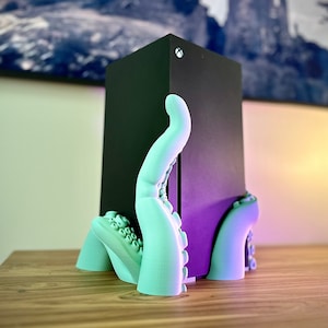 Peut inclure: Un support d'octopus imprimé en 3D en turquoise tenant une console de jeu noire. La console a un logo blanc sur le dessus.