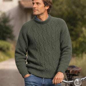 Pode incluir: Uma camisola verde-azeitona de manga comprida com um padrão de malha trançada na frente, gola redonda canelada e punhos canelados. A camisola é usada com calças de ganga azuis, exibindo uma construção em malha texturizada e um design clássico.