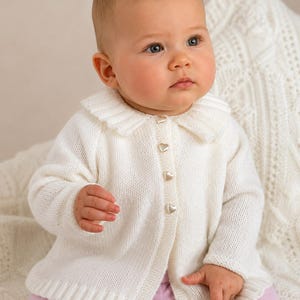 May include: A white long-sleeved knit cardigan featuring a rounded collar and a front button closure with four small, metallic heart-shaped buttons. The garment is constructed with a fine, smooth knit texture and has a clean, solid white colour.