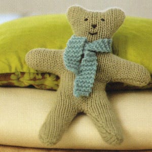 May include: A handmade knitted teddy bear in a light brown color, wearing a blue scarf. The bear has a simple stitched face and outstretched arms. The soft toy is resting on a light-colored surface.
