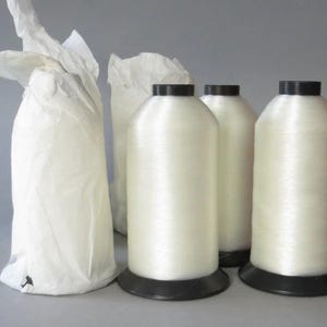 May include: Several spools of white thread with black bases are displayed, alongside two white paper bags. The thread spools are cylindrical and appear to be made of a shiny, smooth material. The bags are crumpled and appear to be empty.