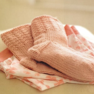 May include: A pair of light pink knitted socks with textured detailing on the cuffs and heels. The socks are resting on a folded piece of fabric with a pink floral pattern. The socks appear soft and cozy.