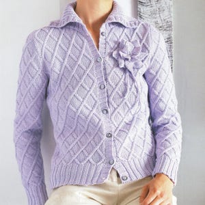 May include: A light purple, button-down cardigan with a diamond pattern and a flower embellishment. The long-sleeved sweater has a collared neckline and is made of a soft, knitted material. The cardigan is worn over a pair of beige pants.
