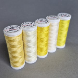 Peut inclure: Six bobines de fil de polyester jaune, chacune étiquetée "TALIA 120" et "200m". Les couleurs de fil varient du jaune pâle au jaune vif, avec des bobines et des couvercles blancs. Les bobines sont disposées en diagonale sur un fond gris.