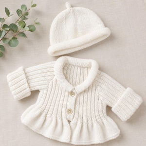 May include: A cream-coloured knitted baby outfit, comprising a jacket with a ruffled hem and a matching cap. The jacket features a collar, button closure, and ribbed cuffs. The set is made of soft, textured yarn.