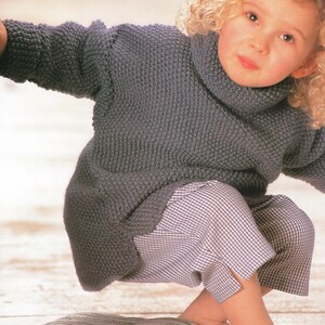 Può includere: Un maglione grigio lavorato a maglia con collo alto e maniche lunghe. Il maglione è abbinato a pantaloni a quadri. Il bambino indossa l'outfit.