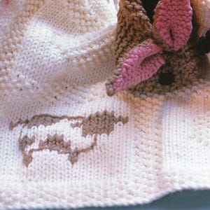 Puede incluir: Primer plano de una manta de bebé de punto color crema con un diseño de oveja marrón y blanco. La manta presenta una cabeza de oveja de punto con orejas rosas y ojos negros. La manta está hecha de hilo suave.
