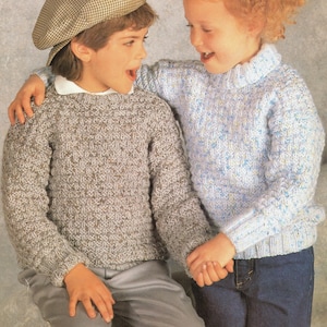 May include: Two children wearing knitted sweaters. One child is wearing a gray sweater with a textured pattern, a white collared shirt, and a plaid cap. The other child is wearing a light blue sweater with a textured pattern and a turtleneck collar. Both children are smiling.