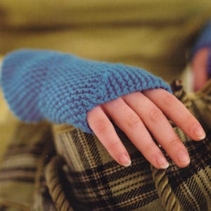 Könnte beinhalten: Nahaufnahme eines gestrickten blauen Fingerhandschuhs. Der Handschuh ist aus strukturiertem Garn gefertigt und hat eine gerippte Manschette. Die Hand ruht auf einem karierten Stoff.