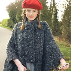 May include: A woman wearing a red beret and a gray knit cape with a matching scarf. The cape has a speckled pattern. The woman has braided hair and is holding a metal object. The background is a blurred outdoor scene.