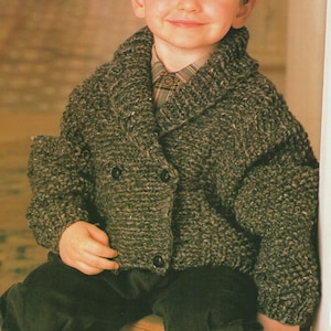 May include: A child wearing a chunky, gray knit jacket with a shawl collar and two dark buttons. The jacket is paired with a plaid shirt and dark corduroy pants. The knitwear appears to be hand-knitted.