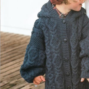 May include: A dark blue hooded cardigan featuring a chunky cable knit pattern with diamond motifs and vertical columns. The garment has long sleeves with ribbed cuffs and a front button closure with dark circular buttons, worn over a plaid shirt.