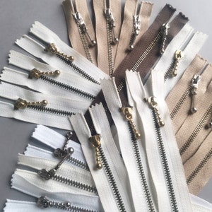 Op de afbeelding: Assortiment van witte, bruine en beige ritsbanden met gouden metalen ritsen. De ritsbanden zijn in een waaier vorm gerangschikt.