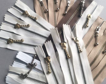 May include: Assortment of white, brown, and beige zipper tapes with gold metal zipper pulls. The zipper tapes are arranged in a fan shape.