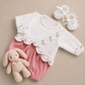 May include: A white knitted long-sleeve top with a scalloped collar featuring small pink floral embroidery, paired with pink corduroy pants, a pair of white knitted booties with ribbon ties, and a small beige plush rabbit toy with a pink ribbon.