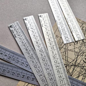 Könnte beinhalten: Eine Sammlung von Metalllinealen mit geätzten Markierungen, die sowohl metrische als auch imperiale Maße anzeigen. Die silberfarbenen Lineale liegen auf einer neutralen Oberfläche, mit Vintage-Schnittmustern im Hintergrund. Die Lineale werden zum Nähen und Zuschneiden verwendet, mit Messungen in cm.