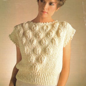 May include: A cream-coloured, short-sleeved knitted top with a textured leaf pattern. The top has a ribbed hem and a simple round neckline. The knitwear has a classic, vintage aesthetic, suitable for casual wear. The overall design is simple and elegant.