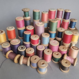 Puede incluir: Una colección de carretes de madera vintage llenos de hilo colorido. Los carretes son de varios tonos de rosa, azul, verde, amarillo y morado. Algunos carretes tienen etiquetas. Los carretes están dispuestos sobre una superficie gris.