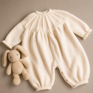 May include: A cream-colored long-sleeve knit romper featuring a ribbed yoke with vertical textured panels, ruffled sleeve cuffs, and a button-down leg closure, accompanied by a small, light brown plush rabbit toy with long ears.
