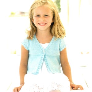 May include: A light blue knitted cardigan with short sleeves and a single button closure at the neckline, worn over a white dress with a textured hem. The cardigan features a ribbed trim along the front opening and sleeve edges.