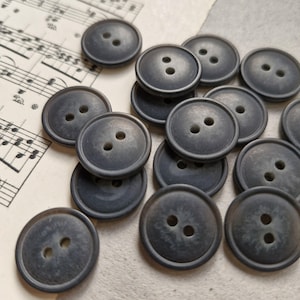 May include: A collection of dark grey buttons with two holes each. The buttons are round and have a slightly textured surface.