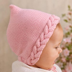 May include: A pale pink knitted baby bonnet with a braided edge. The bonnet features a pointed crown and is made from soft yarn. The baby is wearing a white garment.
