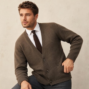 Peut inclure: Un cardigan en tricot marron doté d'une encolure en V, de manches longues et d'une fermeture boutonnée sur le devant avec trois boutons visibles. Le vêtement comprend deux poches plaquées et se porte sur une chemise blanche à col et cravate.