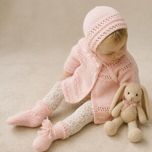 May include: A light pink knitted baby cardigan with a matching bonnet featuring a delicate eyelet pattern and ribbon ties. The set is paired with white lace-patterned tights, light pink knitted booties with tassels, and a beige knitted rabbit toy.