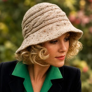 May include: A beige speckled knit cloche hat featuring a textured cable knit pattern around the crown and a wide, slightly downturned brim. The hat is crafted from a flecked yarn that incorporates small brown and dark spots throughout the fabric.