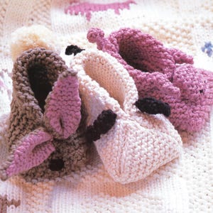 May include: Three knitted bunny slippers in brown, cream, and pink, with black accents. The slippers have large, floppy ears and are arranged on a cream-colored knitted surface. The slippers are designed for babies or toddlers.