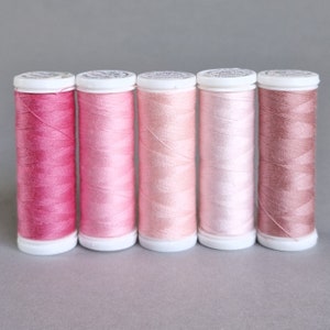 May include: Six spools of pink thread, ranging from a deep pink to a light peach color. The spools are all the same size and have white plastic caps.