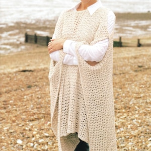 May include: A cream-colored, open-weave crochet poncho with armholes and a long, flowing design. The poncho is worn over a white collared shirt. The image is taken outdoors on a beach.
