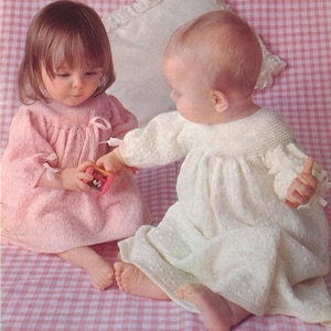 May include: Two babies wearing knitted dresses are seated on a pink and white checkered surface. One baby wears a light pink dress and the other wears a white dress. The baby in pink is holding a small toy. Both dresses have long sleeves.
