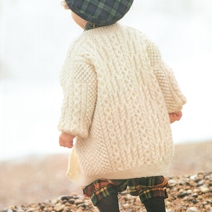 Könnte beinhalten: Ein cremeweißer Zopfmuster-Pullover mit langen Ärmeln und strukturiertem Muster, kombiniert mit einer dunkelgrünen und marineblauen karierten Baskenmütze und passender karierter Shorts. Der Pullover zeigt komplexe vertikale Zopfmuster.