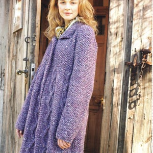 Può includere: Un lungo cappotto viola strutturato con un design a trecce. Il cappotto ha un collo e una chiusura con bottoni. La donna indossa una sciarpa fantasia. Lo sfondo è una porta di legno.