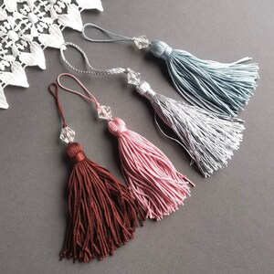 May include: Four tassel keychains with crystal beads. The tassels are in shades of brown, pink, silver, and light blue.