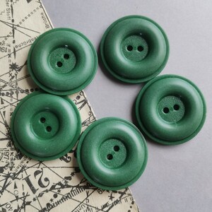 Op de afbeelding: Vijf groene plastic knopen met elk twee gaten. De knopen zijn rond met een verhoogde rand en een licht getextureerd oppervlak.