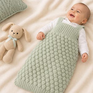 May include: A sage green knitted baby sleep sack featuring a textured bobble stitch pattern on the main body and ribbed detailing at the chest and hem, paired with a crocheted beige rabbit toy with a light green ribbon around its neck.