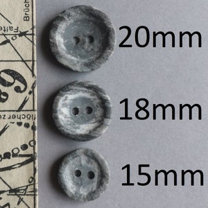 Puede incluir: Tres botones de mármol gris y blanco con dos agujeros cada uno. Los botones tienen un diámetro de 20 mm, 18 mm y 15 mm.