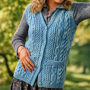 May include: A light blue cable-knit vest with a V-neck, two front pockets, and button closures. The vest features a textured pattern and is worn over a gray long-sleeved shirt. The vest is a cozy, handmade garment.