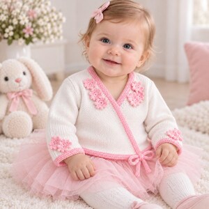 Puede incluir: Un cárdigan cruzado de punto color crema con ribetes rosas, mangas largas y aplicaciones florales rosas en el pecho y los puños. Se combina con una falda de tul rosa de varias capas, medias blancas y un pequeño lazo rosa para el pelo.